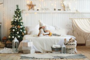 Cozy little sisters are hugging on the bed in white warm sweaters and knitted hats in a bright Christmas interior, the concept of waiting for Christmas and New Year