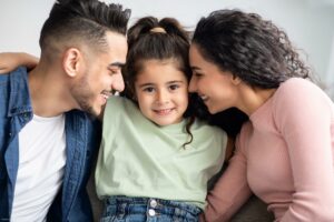 Parental Love. Happy Arabic Parents Cuddling Their Cute Little Daughter At Home