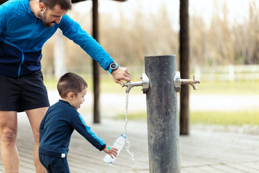 Funny little boy filling his water bottle in public fountain while his father helps him.